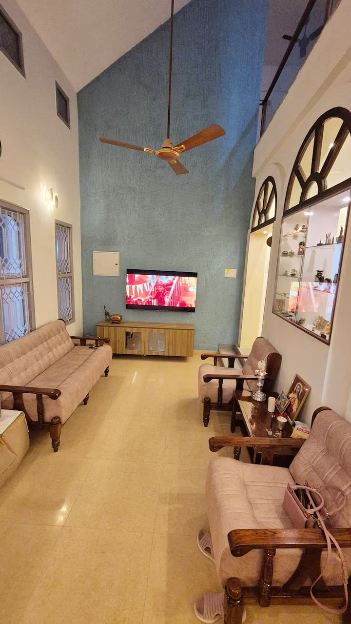 The living room under its double-height ceiling, with a teal feature wall, wall-mounted TV and wooden sofas